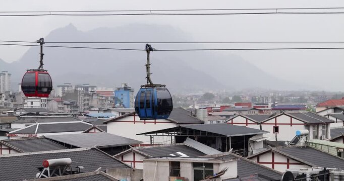 Red and blue cable cars are gliding along a funicular line above the dense rooftops of Chinese city, with misty mountains providing a beautiful scenic background for this public transit system