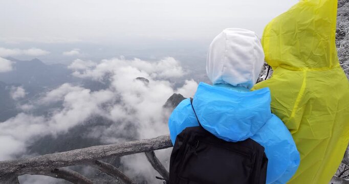 Tourist couple stand ta cliffside viewpoint at Tianmen Mountain National Forest Park, looking down to winter landscape and white clouds. Camera move forward to reveal view they looking to. After night