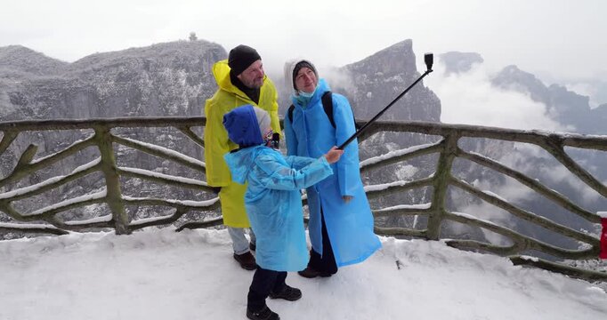 Tourists family in colorful raincoats taking selfie video with action camera on selfie stick on snowy viewing platform, revealing majestic, white landscape of Tianmen Mountain National Forest Park