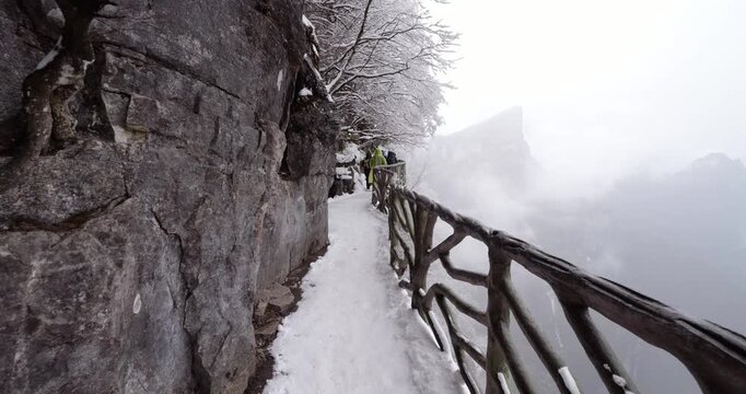 First-person perspective walking along narrow, snow-covered cliffside trail at Tianmen Mountain. Cinematic slow motion captures snow falling from frosted trees onto empty path