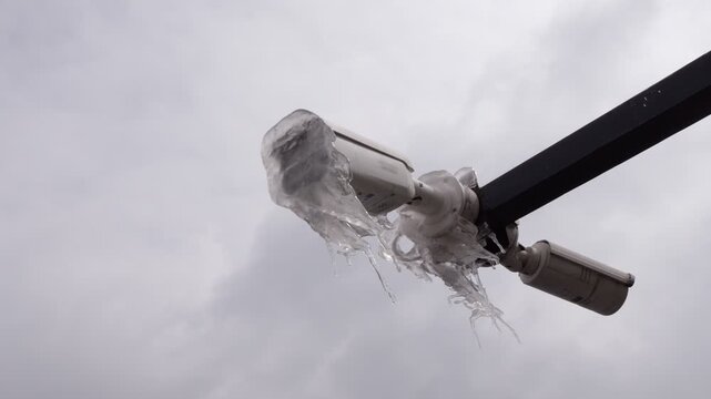 Low-angle parallax shot shows outdoor security camera encased in thick layer of glaze ice and long icicles. Following intense night storm in late March, surveillance equipment is rendered inoperable