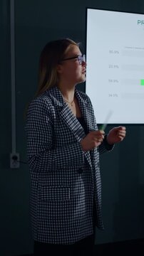 Young professional woman giving analytical business presentation with projected performance chart in modern dark conference room, holding notes while speaking