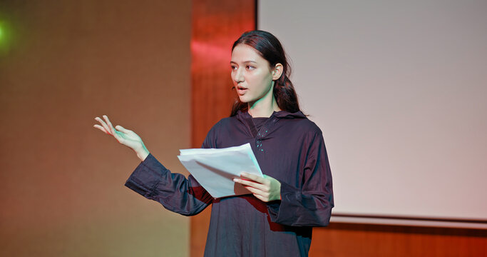 Indian young adult gen z female lady standing wear outfit dress hold script paper do play scene practice indoor hall stage. Teen age girl read story line talk speak day time college campus room place