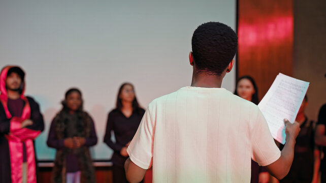 African young adult gen z male man hold scripts paper read play story lines scene standing indoor stage hall. Happy group boy girl do work drama practice day studio campus place