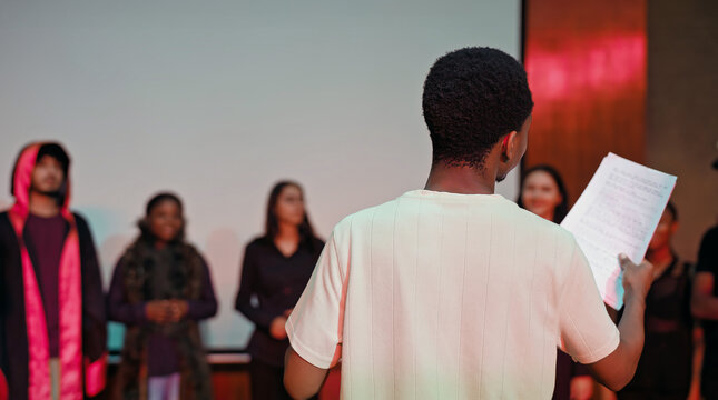 African young adult gen z male man hold scripts paper read play story lines scene standing indoor stage hall. Happy group boy girl do work drama practice day studio campus place