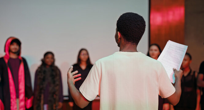 African young adult gen z male man hold scripts paper read play story lines scene standing indoor stage hall. Happy group boy girl do work drama practice day studio campus place