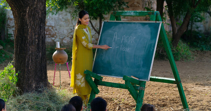 Indian young adult gen z female lady talk speak teach to villager child day under tree rural area. Girl boy group sitting on mat do study look board learn read english at open class field place