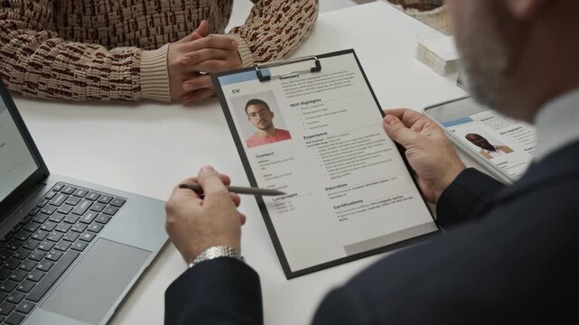 Over the shoulder view of middle-aged HR specialist dressed in formalwear studying gen Z mans CV and asking him questions during job interview in office