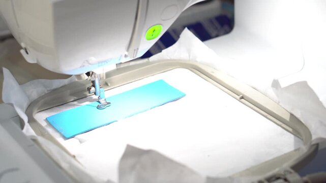 4K Close up of a high-speed computerized embroidery machine stitching a white letter T onto premium blue leather. Precision industrial sewing process and textile manufacturing technology.