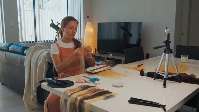 A woman sits at a table preparing to film a video tutorial on hair extension using tools and materials around her.
