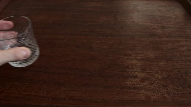 Close up view of hand performing action of setting down or lifting empty crystal glass, highlighting texture of dark wood surface and etched pattern of glass.