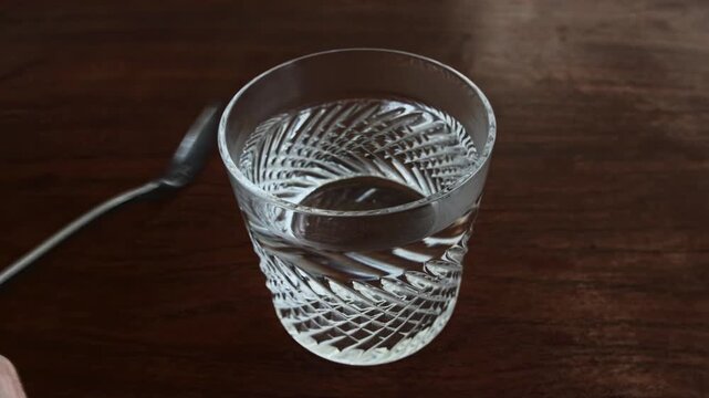 Overhead view clear decorative glass with still water on rich brown wooden table. Metal spoon next to glass, suggesting beverage preparation, consumption. Light reflects on water, highlights glass tra