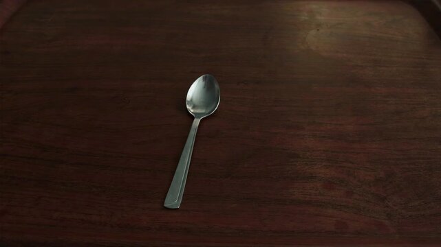 Human hand reaches into frame to pick up shiny metallic spoon from rich brown wood tabletop, then gently sets it back down. Action highlights movement and everyday use of cutlery.