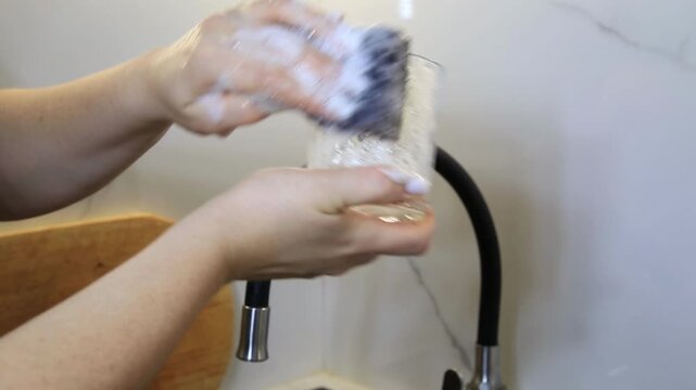 Detail of woman hands performing a routine kitchen chore, scrubbing a clear glass with a sudsy sponge. White foam covers glass as water flows from faucet, emphasizing cleanliness process.