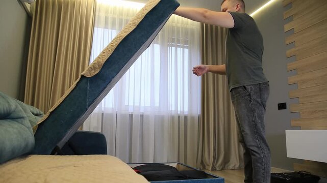 Caucasian man opening storage compartment of sofa by lifting seat cushion. Action reveals hidden space for items, highlighting practical and space-saving furniture solution in modern living room.