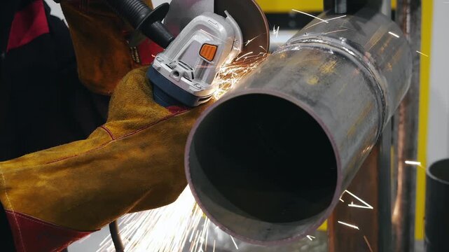 Close up of worker cutting metal pipe with angle grinder producing sparks during metalworking process in industrial workshop.