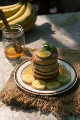 Stack of fluffy homemade banana pancakes topped with fresh slices, mint leaf, and golden honey dripping from a wooden dipper. Rustic breakfast concept on burlap background with natural sunlight. © Chayut