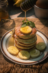 Stack of fluffy homemade banana pancakes topped with fresh slices, mint leaf, and golden honey dripping from a wooden dipper. Rustic breakfast concept on burlap background with natural sunlight. © Chayut