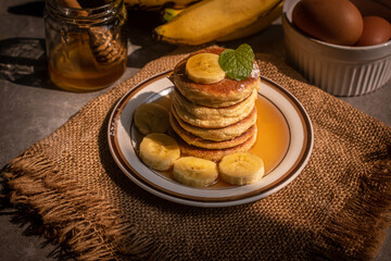 Stack of fluffy homemade banana pancakes topped with fresh slices, mint leaf, and golden honey dripping from a wooden dipper. Rustic breakfast concept on burlap background with natural sunlight. © Chayut