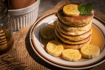 Stack of fluffy homemade banana pancakes topped with fresh slices, mint leaf, and golden honey dripping from a wooden dipper. Rustic breakfast concept on burlap background with natural sunlight. © Chayut
