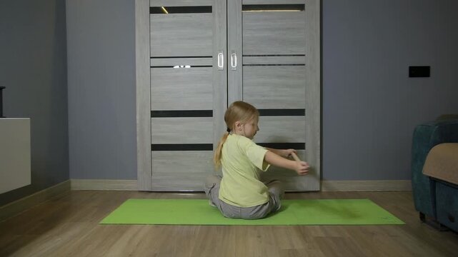 Little girl with blonde ponytail sits cross-legged on green fitness mat, practicing arm extension and retraction. Exercise promotes shoulder and back flexibility indoors on wooden floor near gray wall