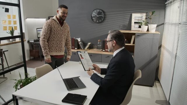 Zoom in shot of gen Z jobseeker showing up in modern office, handing application form to mature HR manager who asking him to take seat at desk and beginning interview after looking at paper
