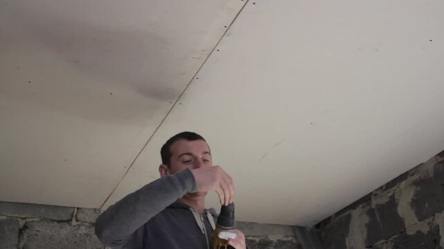 Installing a plasterboard ceiling using an electric drill, Master installs plasterboard sheets on ceiling indoors