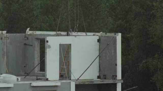 Assembly of modern residential building using heavy machinery and precast concrete elements. Crane lifts large wall panel into position at construction site. Wooden ladder against unfinished structure