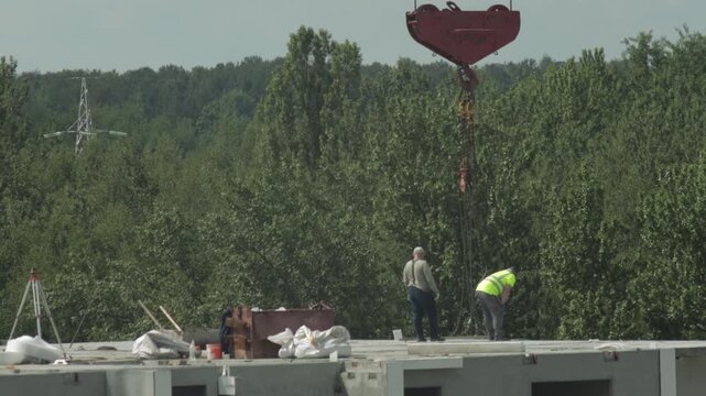 Two construction workers perform duties on concrete building rooftop under overcast sky, large red crane hook poised above. Lush forest line and distant power tower visible, indicating industrial deve