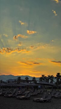 Morning light illuminating empty beach of resort area, featuring organized lines of sunbeds and thatched parasols on dark sand. Silhouetted palm trees and mountain range against dramatic cloud formati