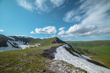 Scenic sunlit view along grassy ridge with white snow cornice to green hill top with rocky sheer crags in bright sun under lush clouds in blue sky. Vivid alpine scenery in high mountains in sunny day. © Daniil