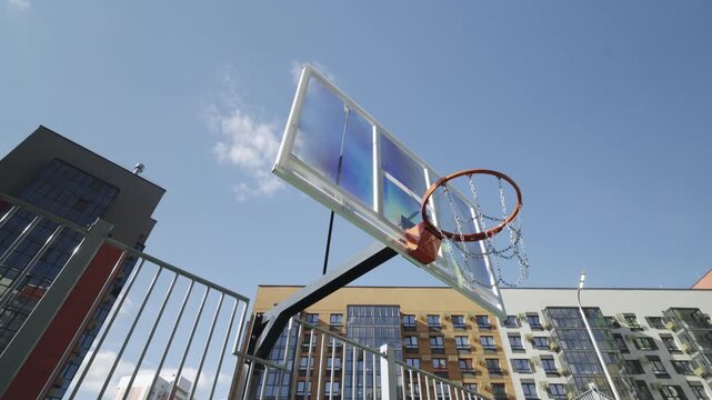 Basketball hoop with transparent backboard shown against blue sky and residential buildings. omposition emphasizes urban atmosphere