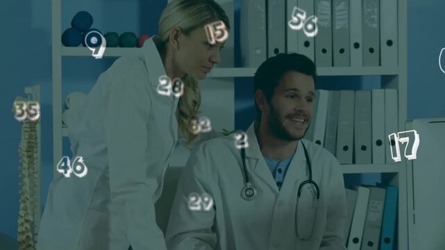 Seated Dr reaching as female Dr handing blue file, viewing medical monitor with numeric overlay