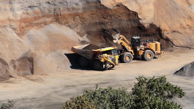 Dump truck parked beside wheel loader as fill and aggregate is loaded for transport, aerial pullback