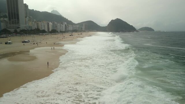Breaking waves and sand at Copacabana beach on winter towards Pedra do Leme. Morning fog present as people go in the water. Shot on 4K Apple ProRes 422. Wide forward aerial over sand and water.