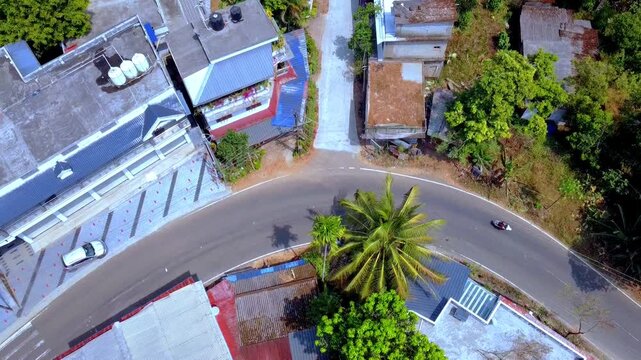 High-angle drone shot of a winding asphalt road in a lush tropical village. Features scooters and cars navigating a sharp bend surrounded by colorful houses and palm trees.