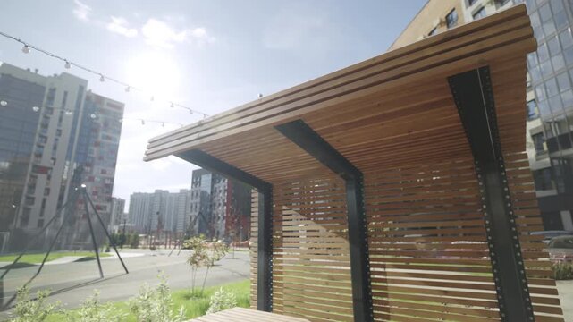 Sunlight illuminates eco friendly wooden pavilion with benches and table. Warm natural materials create harmonious courtyard atmosphere