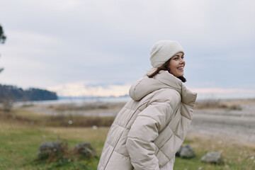 Woman in a warm beige coat and knit beanie enjoying a windy coastal landscape, smiling as she faces the sea with a calm, expansive sky.