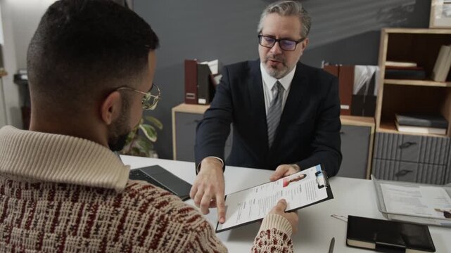 Zoom out over the shoulder shot of mature recruiter in formalwear asking gen Z candidate to sign application form after interviewing him at office desk
