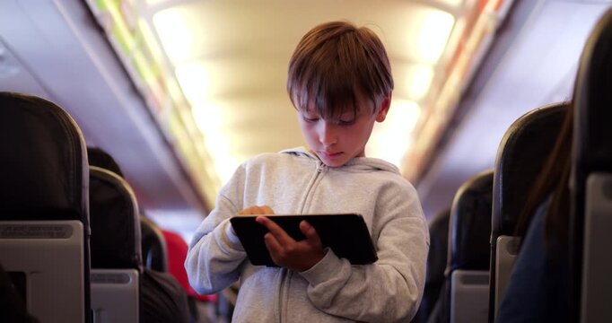 Young boy standing in aircraft aisle while deeply focused on tablet for creative apps or entertainment. Teenager passenger having grown bored of sitting, he finds new way to stay active and engaged
