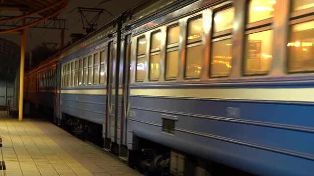 Nighttime scene of a train arriving and moving at the station. Lights illuminate the platform and railway tracks. Urban transportation and city commute atmosphere.