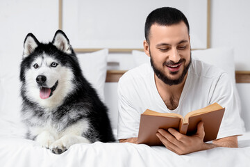 Handsome young man lying on bed and reading book with cute husky dog in bedroom, closeup © Pixel-Shot