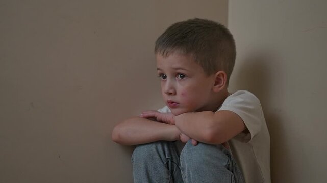 Boy sits in corner wrapped in quiet stillness. Isolation surrounds his posture. Loneliness seen in child expression. Isolation repeats in silence. Loneliness and quiet define isolation moment.