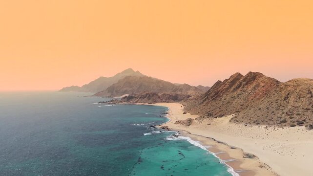 Aerial view of the coastline featuring rugged mountains, sandy beach, and turquoise ocean water under a hazy orange sky, Socotra, Yemen.