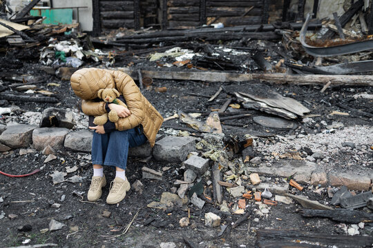 a person sitting amidst the charred remains of a destroyed building. Clad in a tan puffer jacket with the hood pulled down, they are hunched over, tightly clutching a small teddy bear to their chest. 