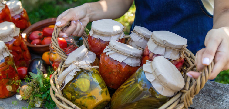 Woman holding wicker basket with jars of preserved vegetables on wooden table
