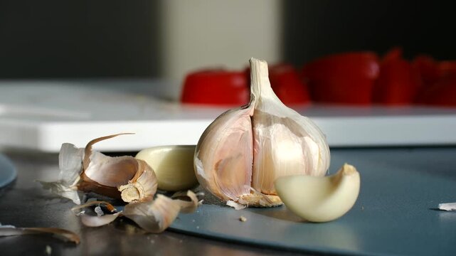 Fresh organic garlic bulb with peeled cloves and skin scattered on a kitchen countertop, with diced red peppers blurred in the background, ready for preparing a healthy meal