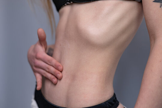 A close-up studio photograph of a young woman&rsquo;s slim torso, waist, and hips against a neutral grey background. The woman, with her head out of frame, has her long brownish hair hanging down on the lef