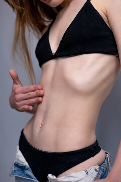 A close-up studio photograph of a young woman&rsquo;s slim torso, waist, and hips against a neutral grey background. The woman, with her head out of frame, has her long brownish hair hanging down on the lef