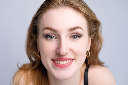 close-up portrait of a smiling young Caucasian woman with light brown hair and green eyes. She features natural-looking makeup, defined eyebrows, and subtle freckles across her nose and cheeks. Her ex